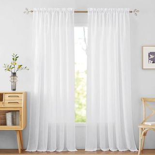 A pair of sheer white linen curtains hanging from a curtain rod against a window inset into a white wall. 