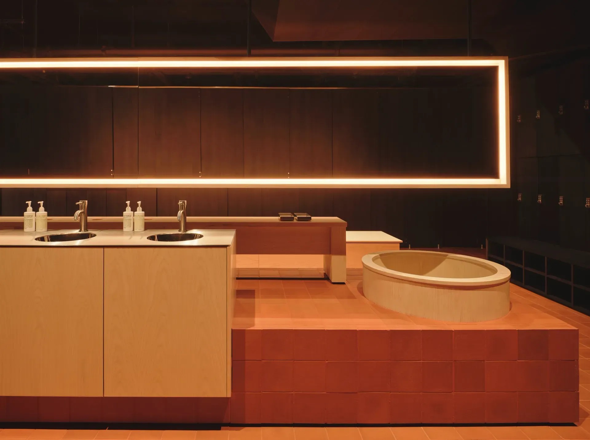 A bathroom decorated with rich red, clay tiles, sculptural neon lighting in a rectangular shape, wooden basins topped with chrome detailing, and wooden storage.