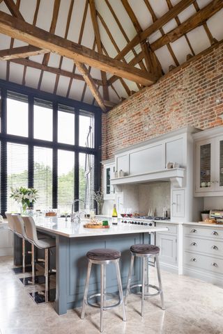 White kitchen with blue gray island, floor to ceiling windows and exposed brick and timber architecture.