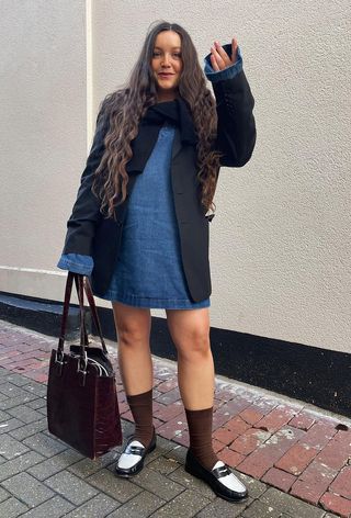 A woman's jean dress outfit with a denim minidress layered underneath black blazer and cardigan and styled with brown socks and two-tone loafers.
