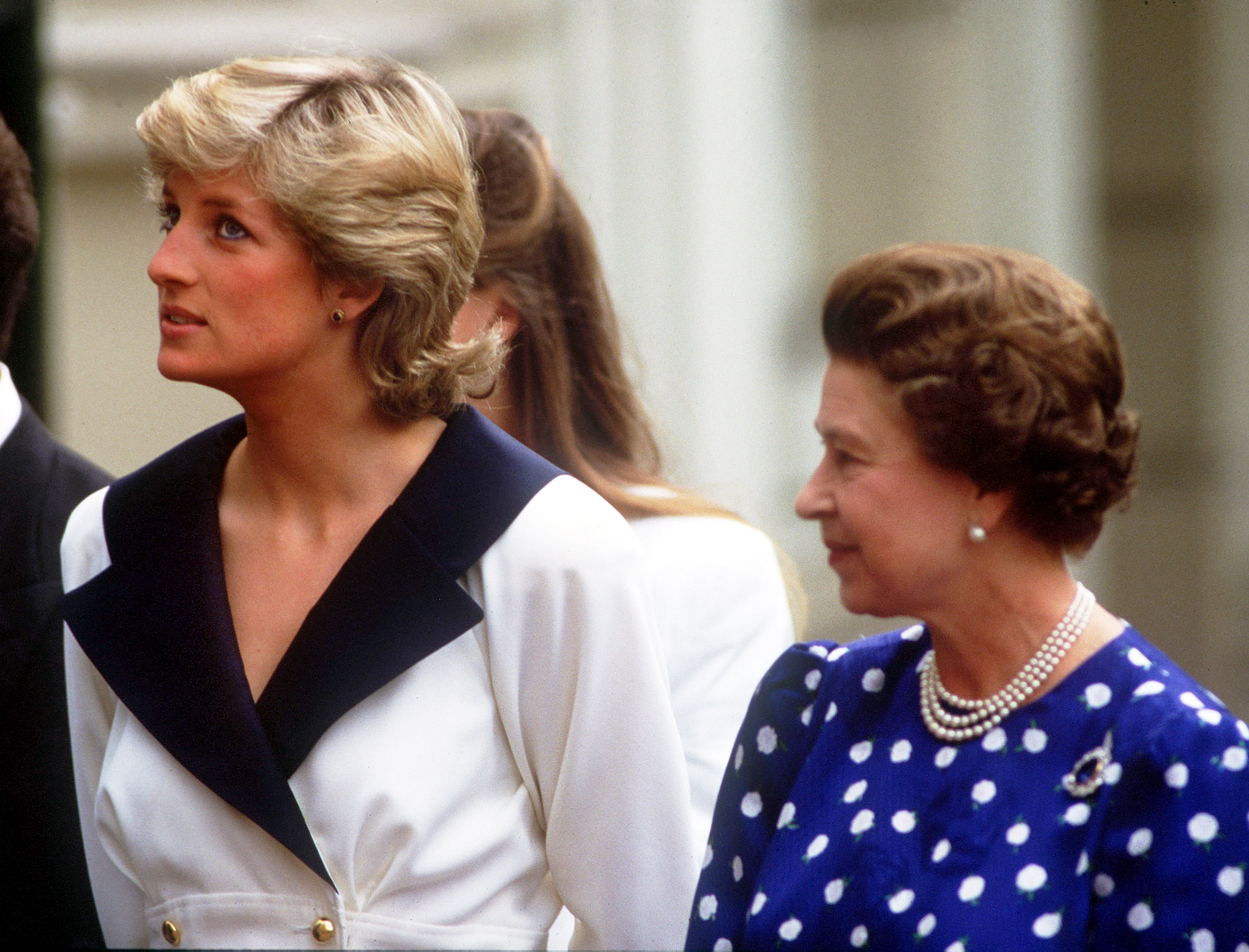 Princess Diana wears a tartan dress with a white collar and stands between Queen Elizabeth II, who wears a light pink coat, and King Charles