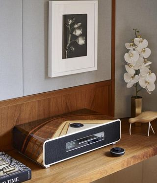A shelf with an audio system (shades of brown case and black face) , bronze vase with white tulips on the right, and a peek of a hard back book on the left. A white framed picture of a flower on the grey wall above the shelf.