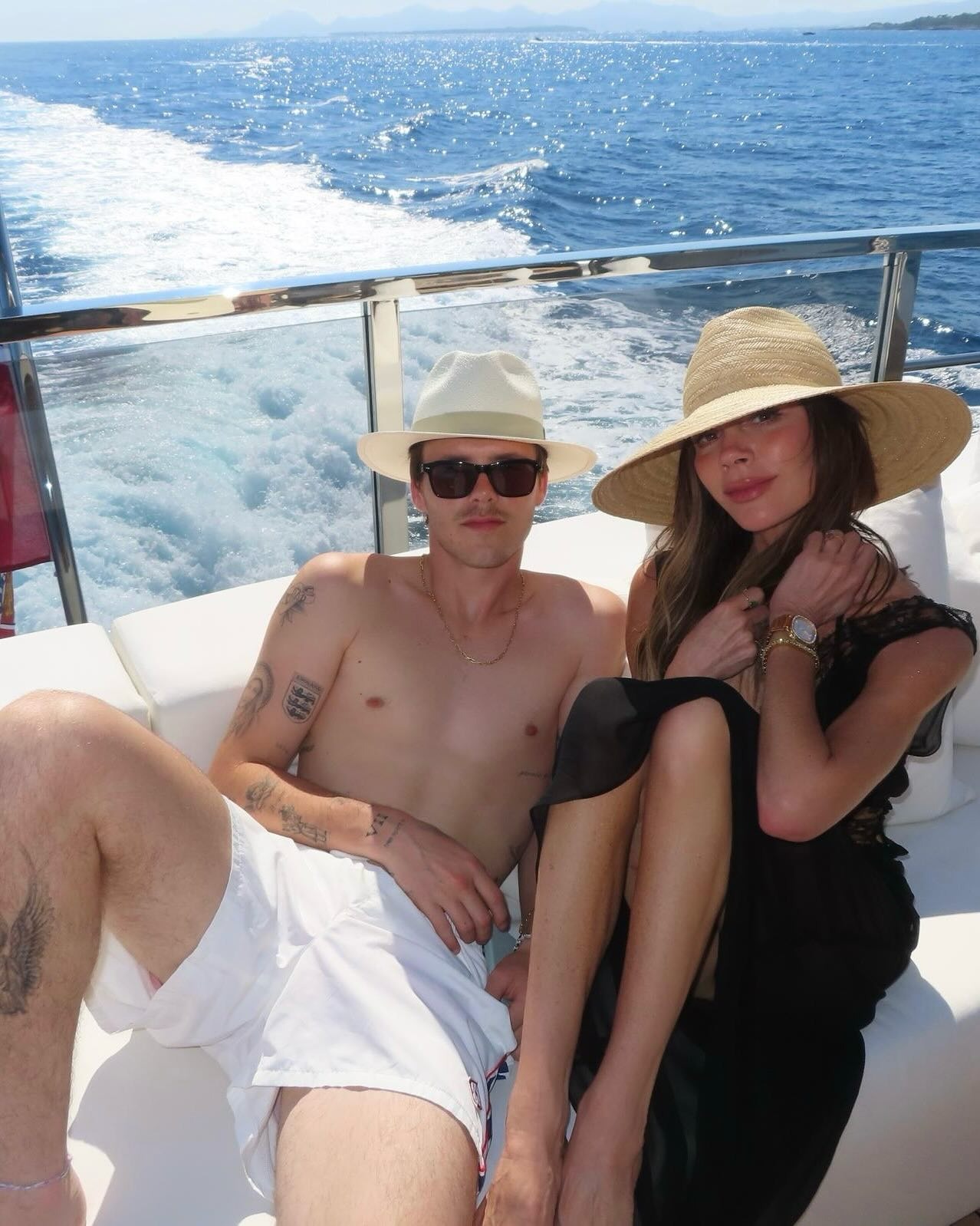 Victoria Beckham sits on a boat wearing a lace trim black dress with an oversized straw hat.