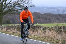 Man riding towards the camera on a green road bike wearing an orange jacket and black helmet.