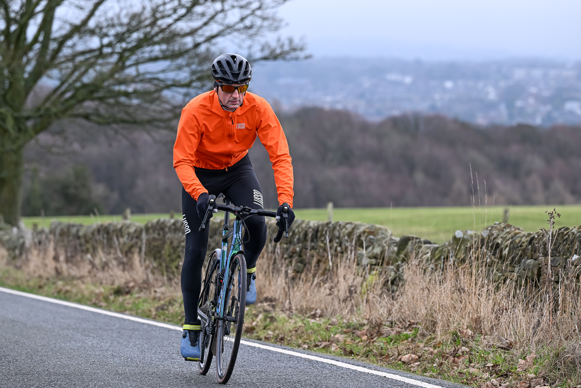 Man riding towards the camera on a green road bike wearing an orange jacket and black helmet.