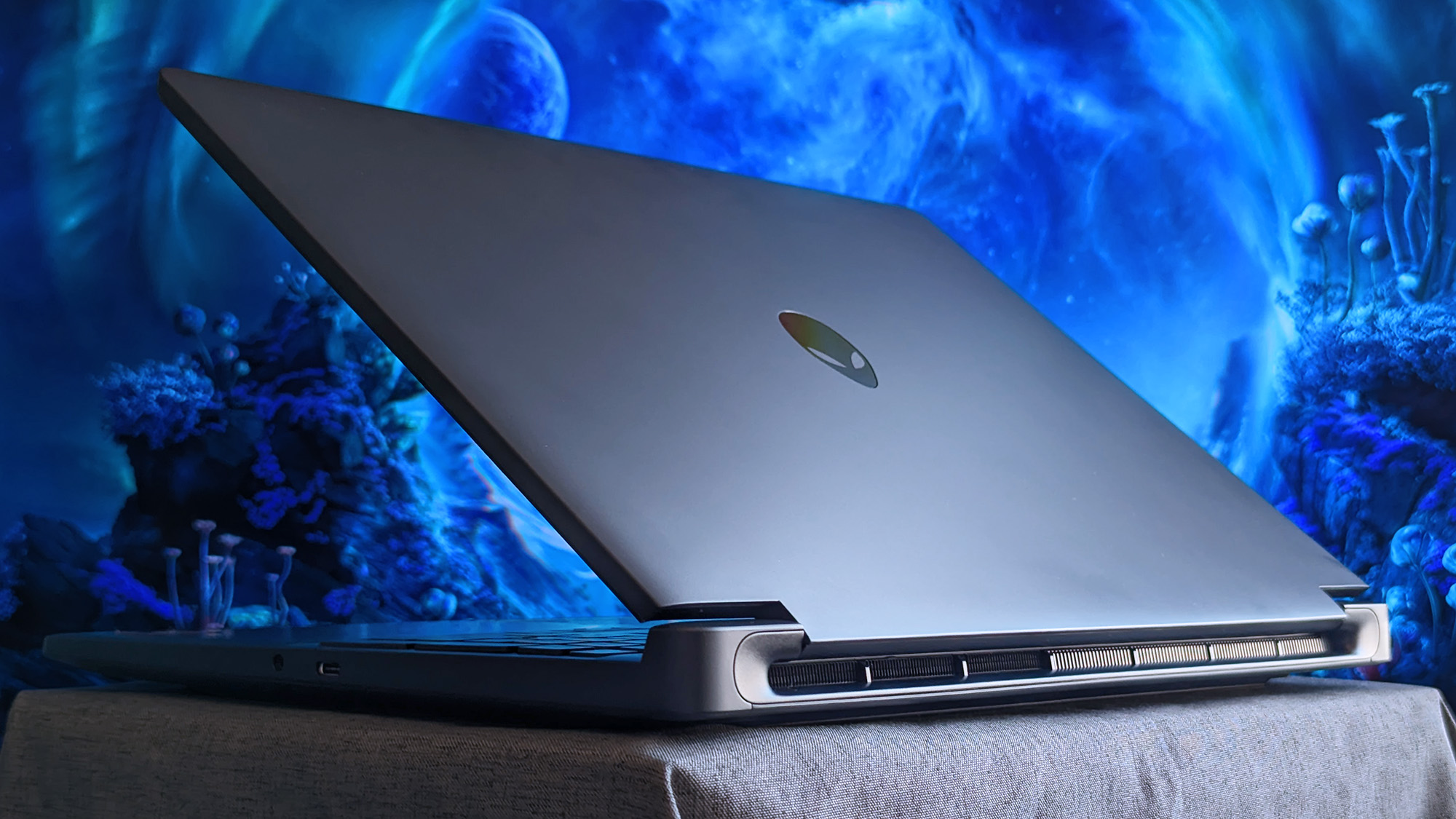 A partially open Alienware 15 laptop rests on fabric, showing its metallic lid with the Alienware logo and rear cooling vents, set against a glowing blue sci&amp;#8209;fi background.