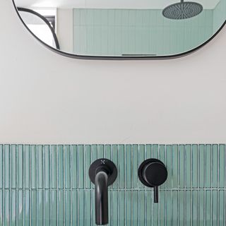 A close-up of matt black bathroom tap accessories fitted on teal mosaic tiles with soft rectangle mirror in background displaying showerhead fixture in reflection