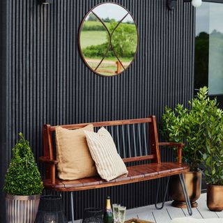 garden bench in front of black wooden cladding wall, metallic planters, yellow cushions, circular metal framed garden mirror, lanterns