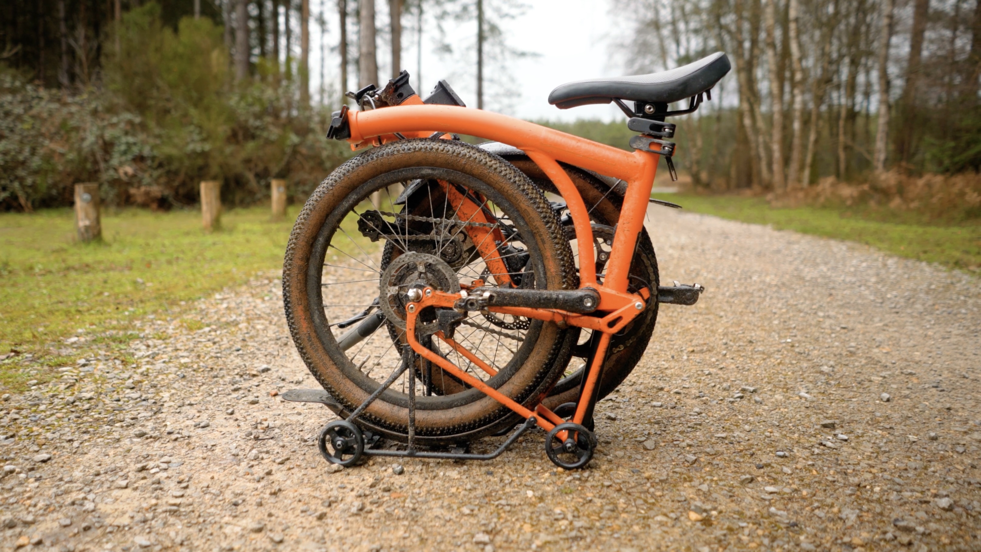 An orange Brompton covered in mud on a light gravel track