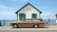 A wood-panelled station wagon is parked outside a small white cottage with green trim and a Cosmos sign, the sea stretching out behind under a pale blue sky.