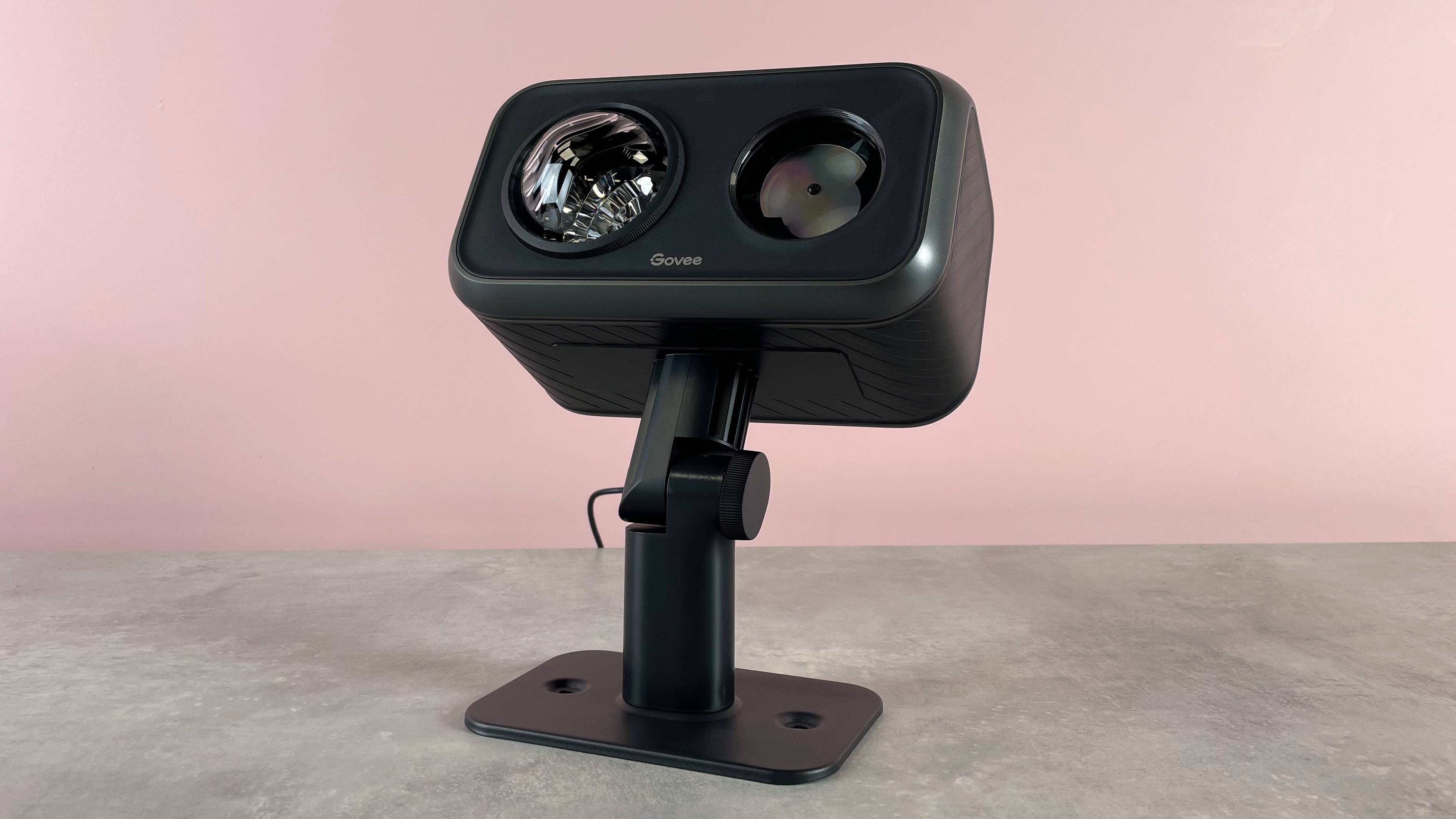 The Govee Outdoor Projector Light is tilted upward and sitting on a grey stone surface against a pink background.