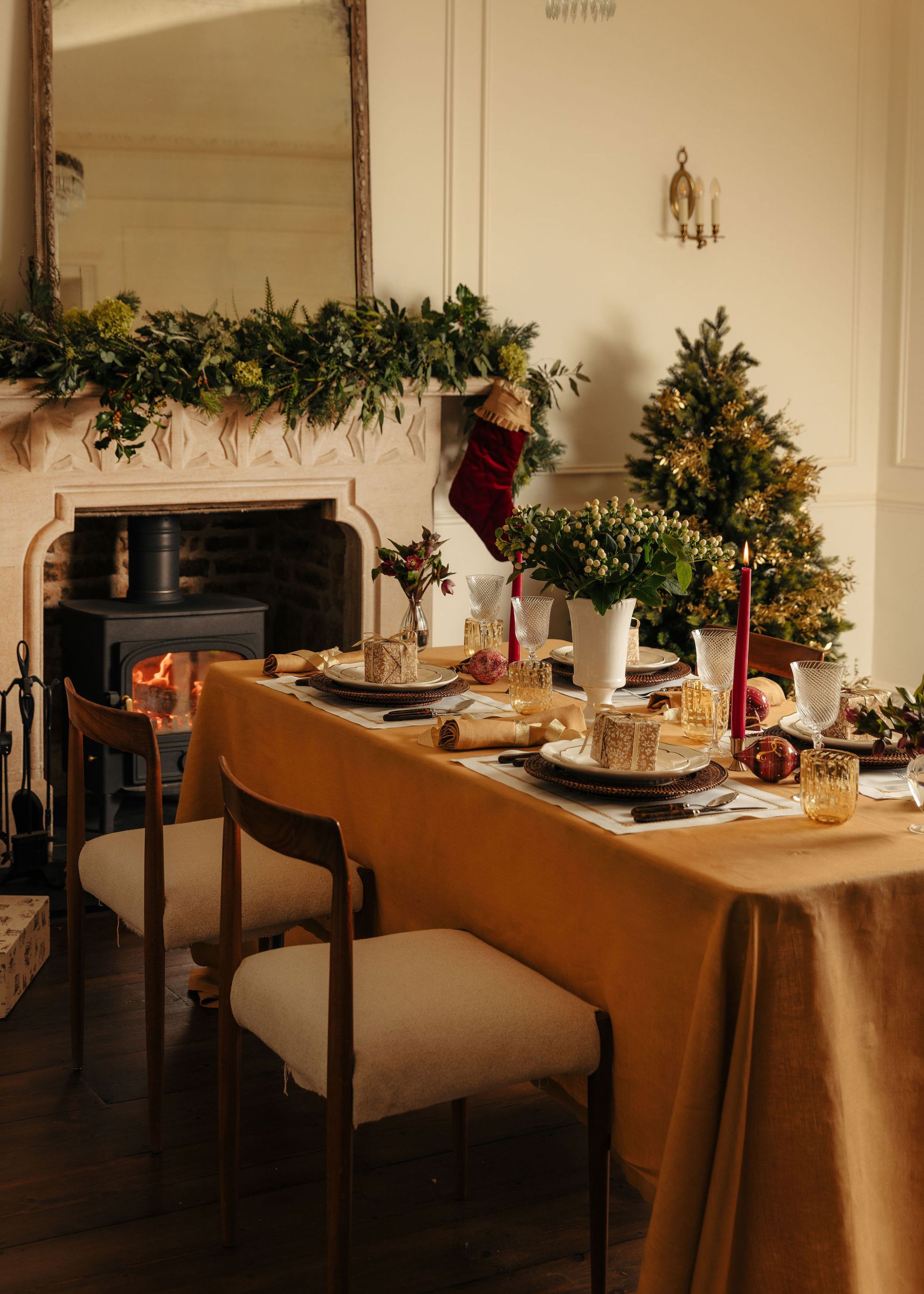 A Christmas tablescape with a tobacco-colored tablecloth, red taper candles, and a fireplace in the corner with a garland.