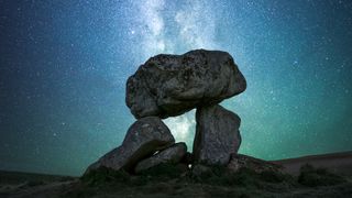 two stones prop up a large upper stone in the foreground. the milky way intersects with the stones in the background. the night sky and stars shine around the rest of the image
