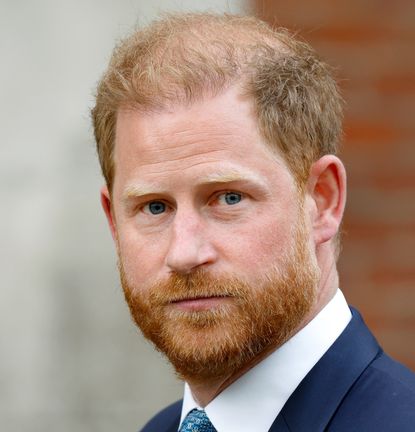 A headshot of Prince Harry wearing a suit and tie in front of a brick wall