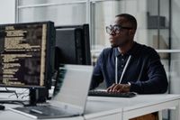 IT worker with glasses sitting at desk looking at computer screen.