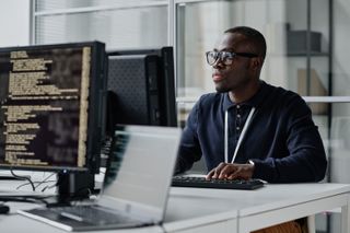 IT worker with glasses sitting at desk looking at computer screen.