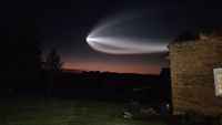 a white streak illuminated in the sky flies over a dark landscape and a brick house. 