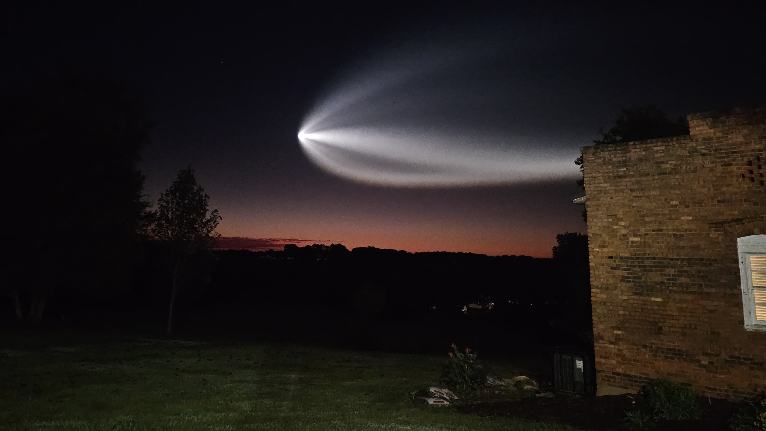 a white streak illuminated in the sky flies over a dark landscape and a brick house. 