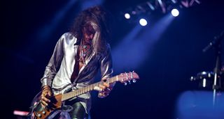 Joe Perry wears a silver shirt and plays his see-through Dan Armstrong live onstage.