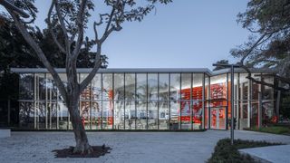 Exterior of Wutopia Lab's, tall glass panels frame the window and entrance door to the building, trees in the white paved pathway, border shrubs and trees in the background, blue dusky sky