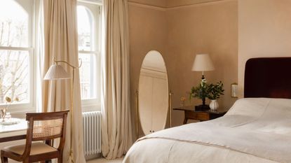 Bedroom with plaster pink walls, dark red velvet bed, linen bedding, round floor mirror, vintage desk with rattan chair, floor lamp and large arched windows