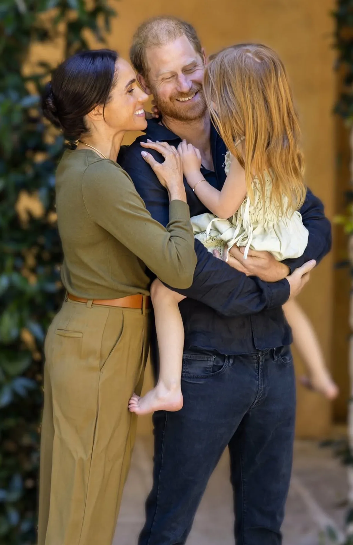 Prince Harry holding Princess Lilibet with Meghan Markle smiling at her