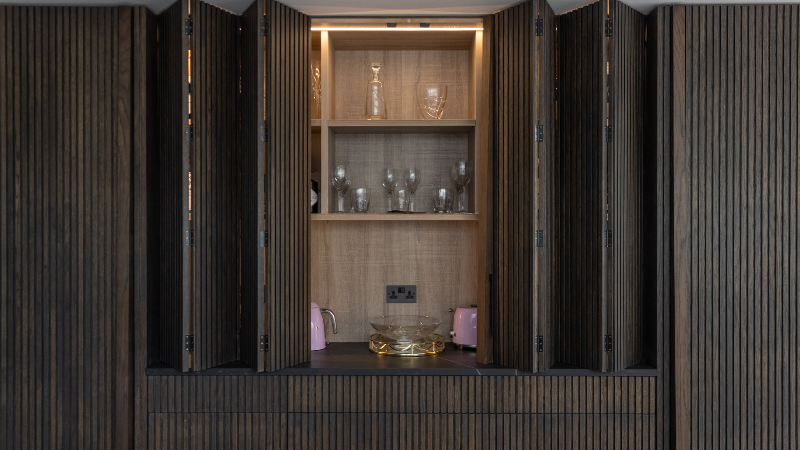 led lighting strip behind a wooden kitchen bifolding door with open shelving and pink smeg appliances within