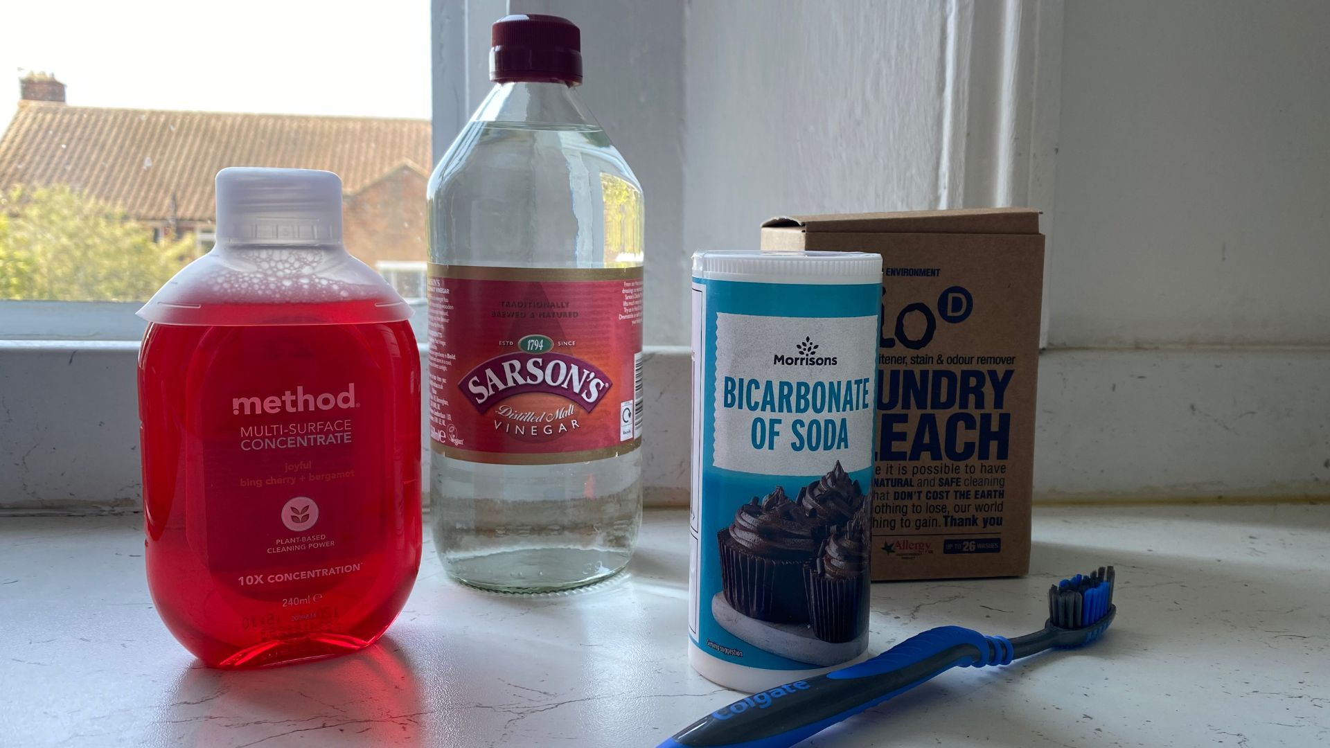 Surface cleaner, white vinegar, baking soda, laundry bleach and a toothbrush photographed in a line on a kitchen counter for an article on how to clean grout