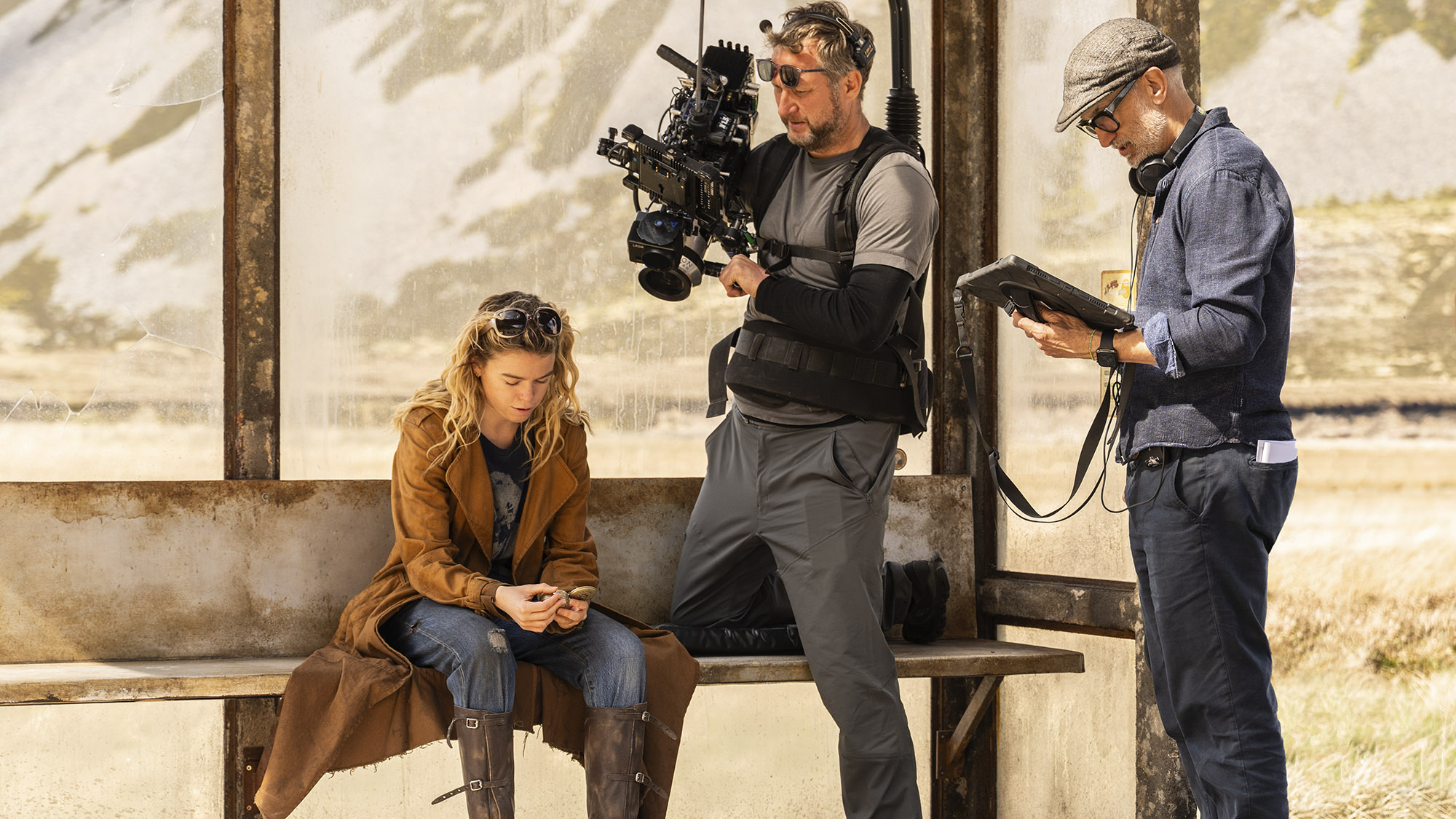 Supergirl (Milly Alcock) sits in a bus shelter while a cameraman and director Craig Gillespie look on.