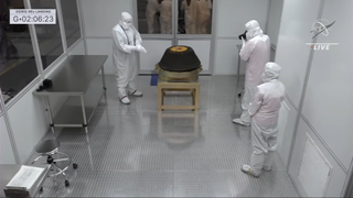 The capsule is seen in the clean room with scientists wearing white outfits gathered around.