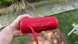 A red JBL Flip 6 held in front of a patch of grass and concrete path. The speaker is seen from behind, so its ports and carry strap are visible.