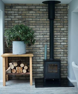 alcove with brick slip tiled wall behind black freestanding log burner, wooden table to side with logs and green plant in grey pot