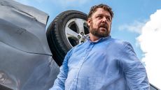 Russell Crowe as Tom Cooper looking unkempt next to an overturned car in Unhinged.