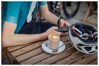 A glass of latte on a table with a cyclist looking at their phone in the background