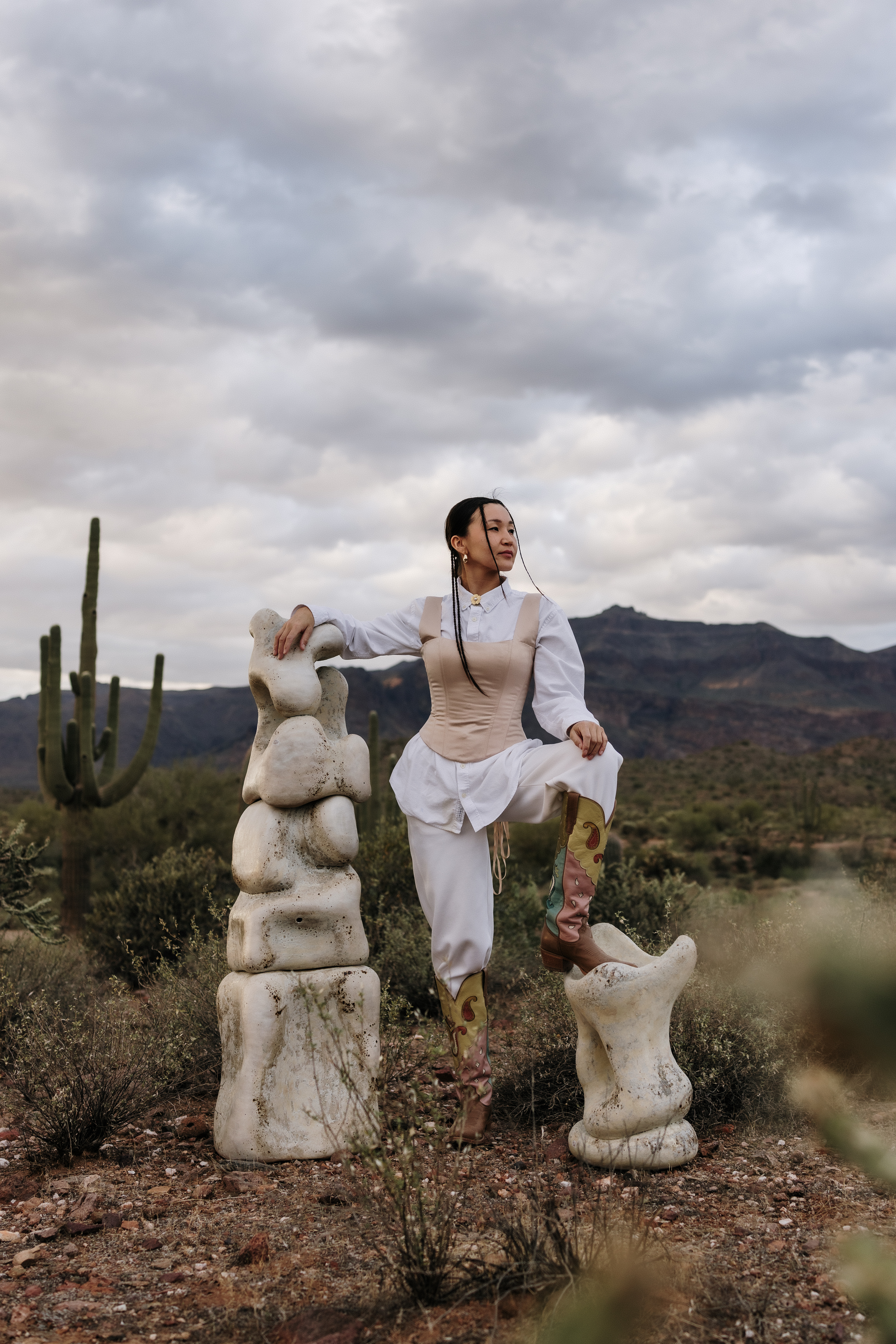 A young woman dressed in white clothing with a beige corsette stares into the distance while standing in an American desert dotted with cacti next to two totemic sculptures.