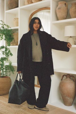 A woman wearing a black peacoat over a green T-shirt styled with black trousers, suede clogs, and a black tote bag.