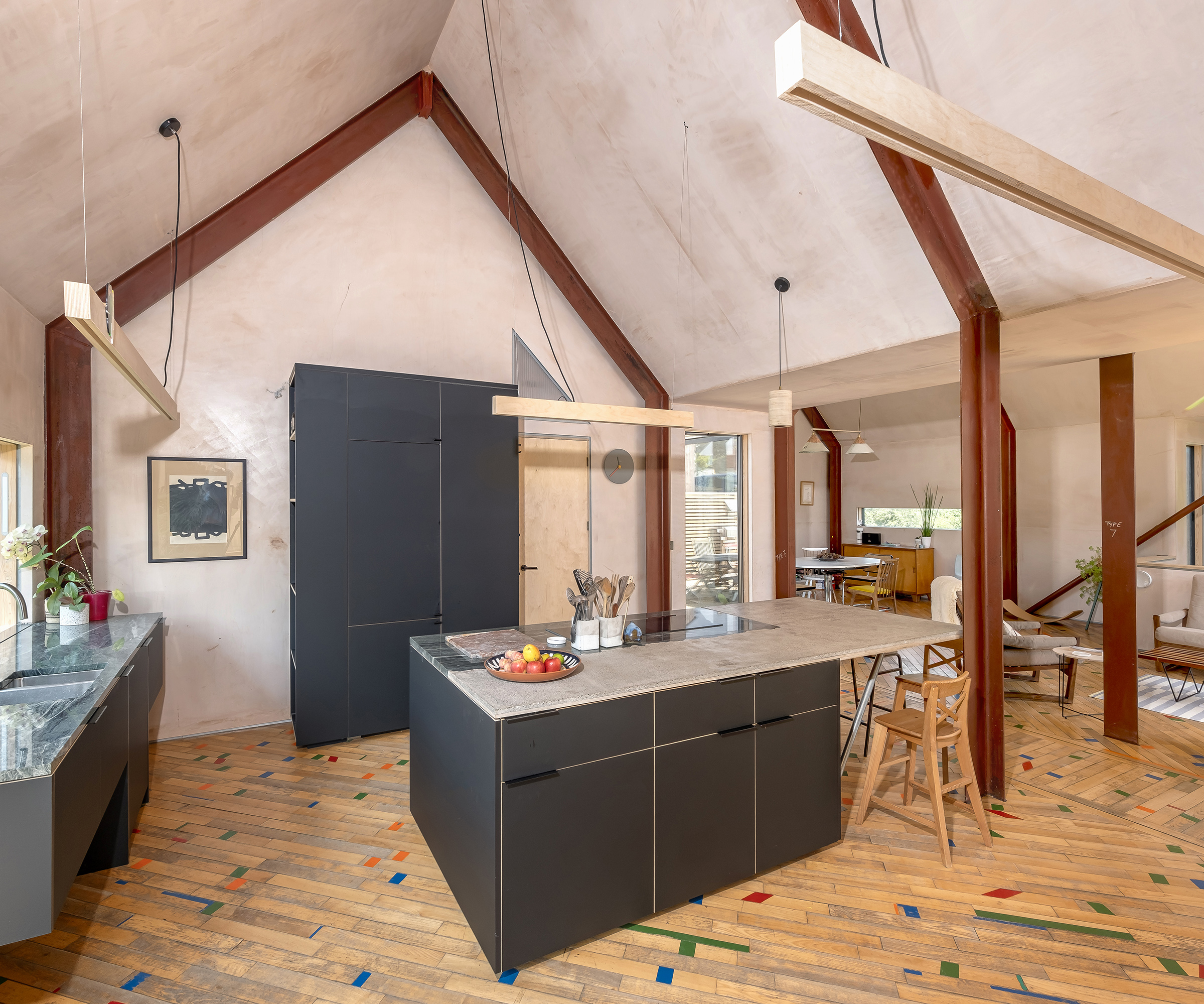 A modern kitchen with dark blue/charcoal cabinets and light surfaces. The ceilings are pitched with exposed beams. Walls are in a plaster colour and the flooring is wood