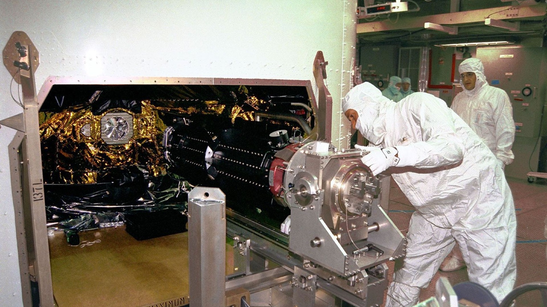 يقوم أشخاص يرتدون بدلات بيضاء نظيفة بتحميل أسطوانة معدنية في صندوق معدني كبير مغطى بالأسلاك