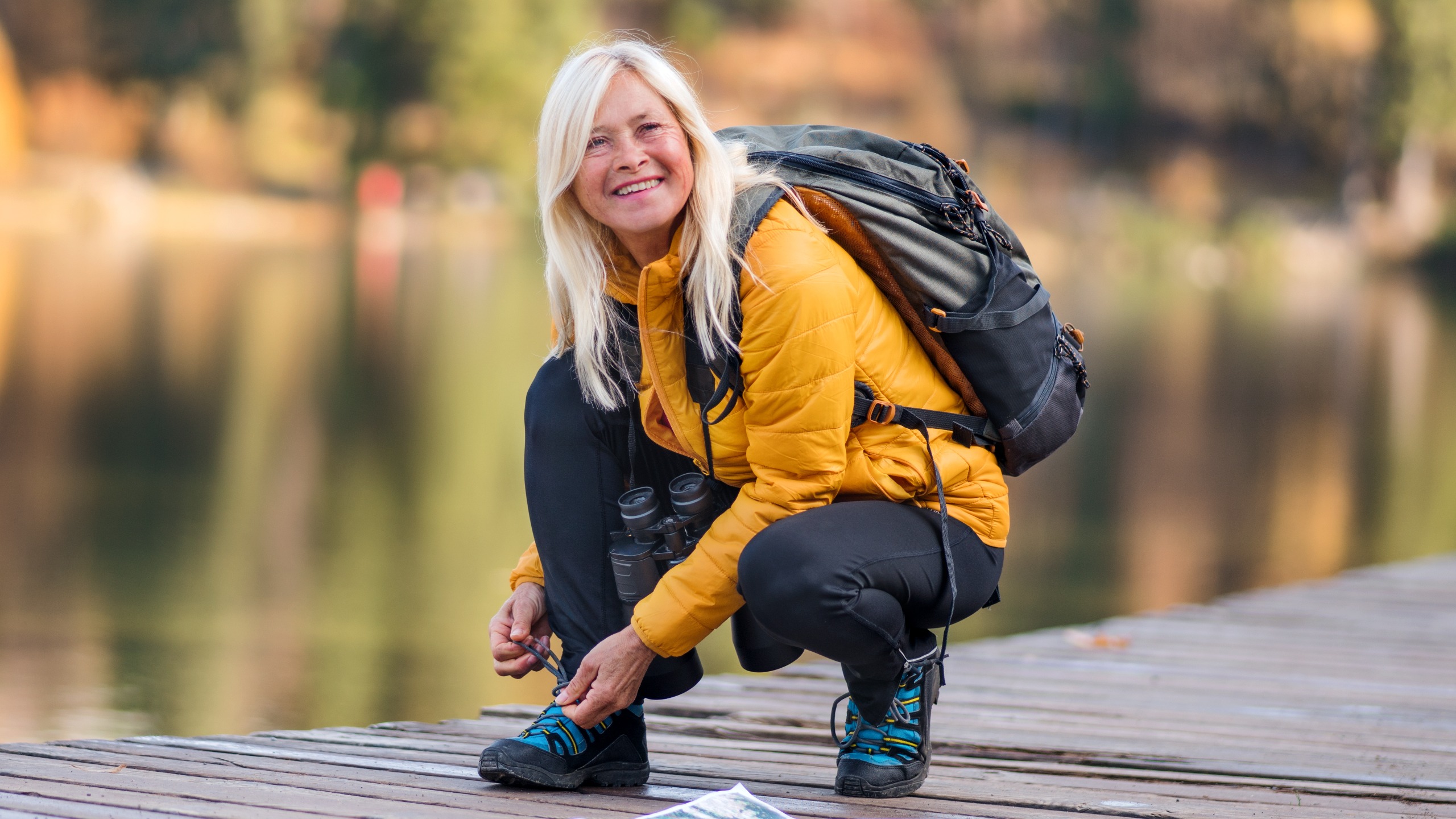 A woman in hiking apparel bends down to tie her shoe on a pier across a lake.