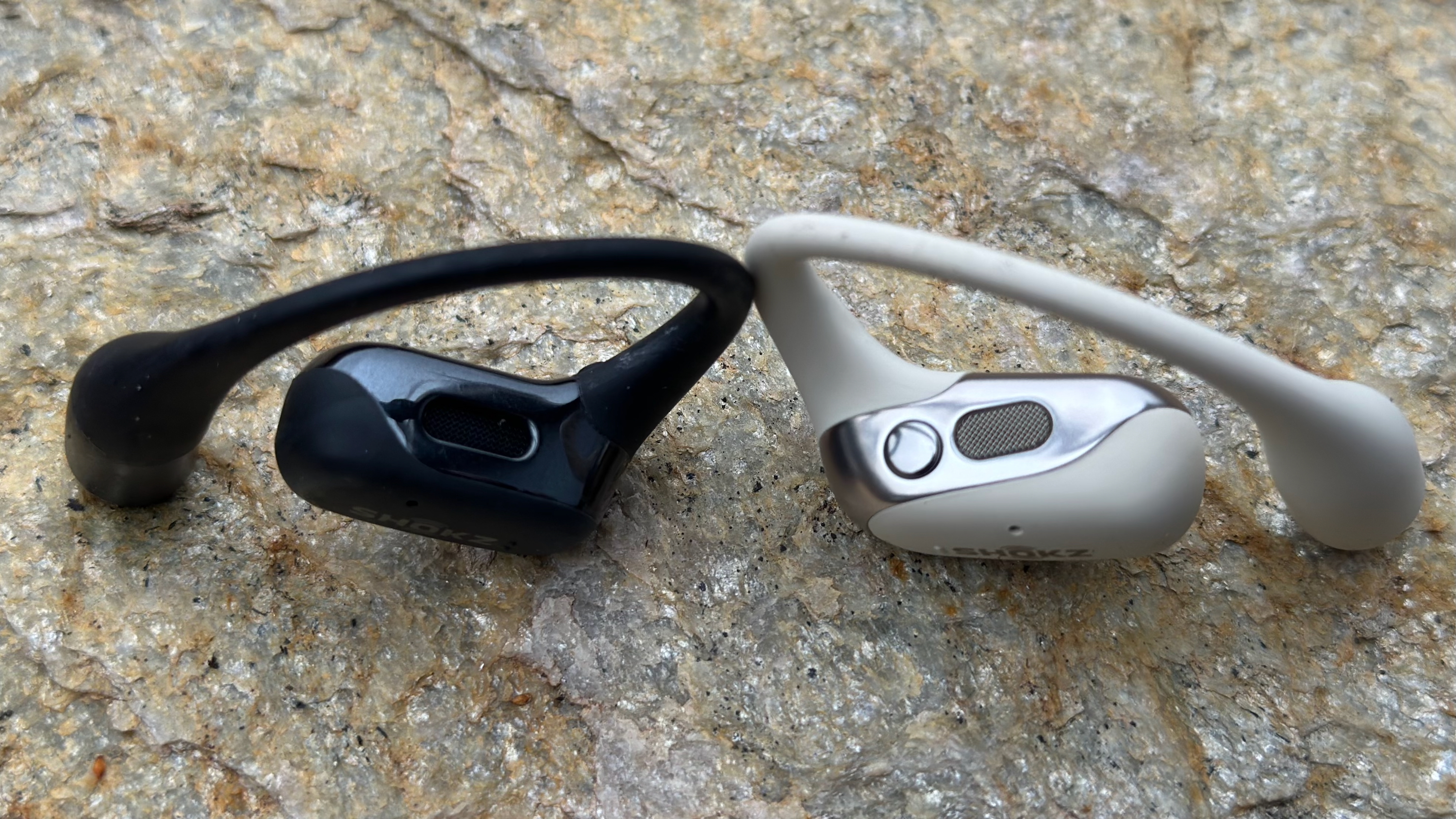 The black Shokz OpenFit and white Shokz OpenFit 2 sitting on a rock upright, the comparison emphasizing the new button on the 2nd-gen model.