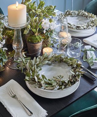 A place setting for Christmas with white candles, glass candlestick, silver green leaf wreaths and pots of greenery, ivy, and moss