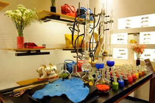 Scientific flasks filled with bright colours displayed on a black table beside shelves of handbags and glasses