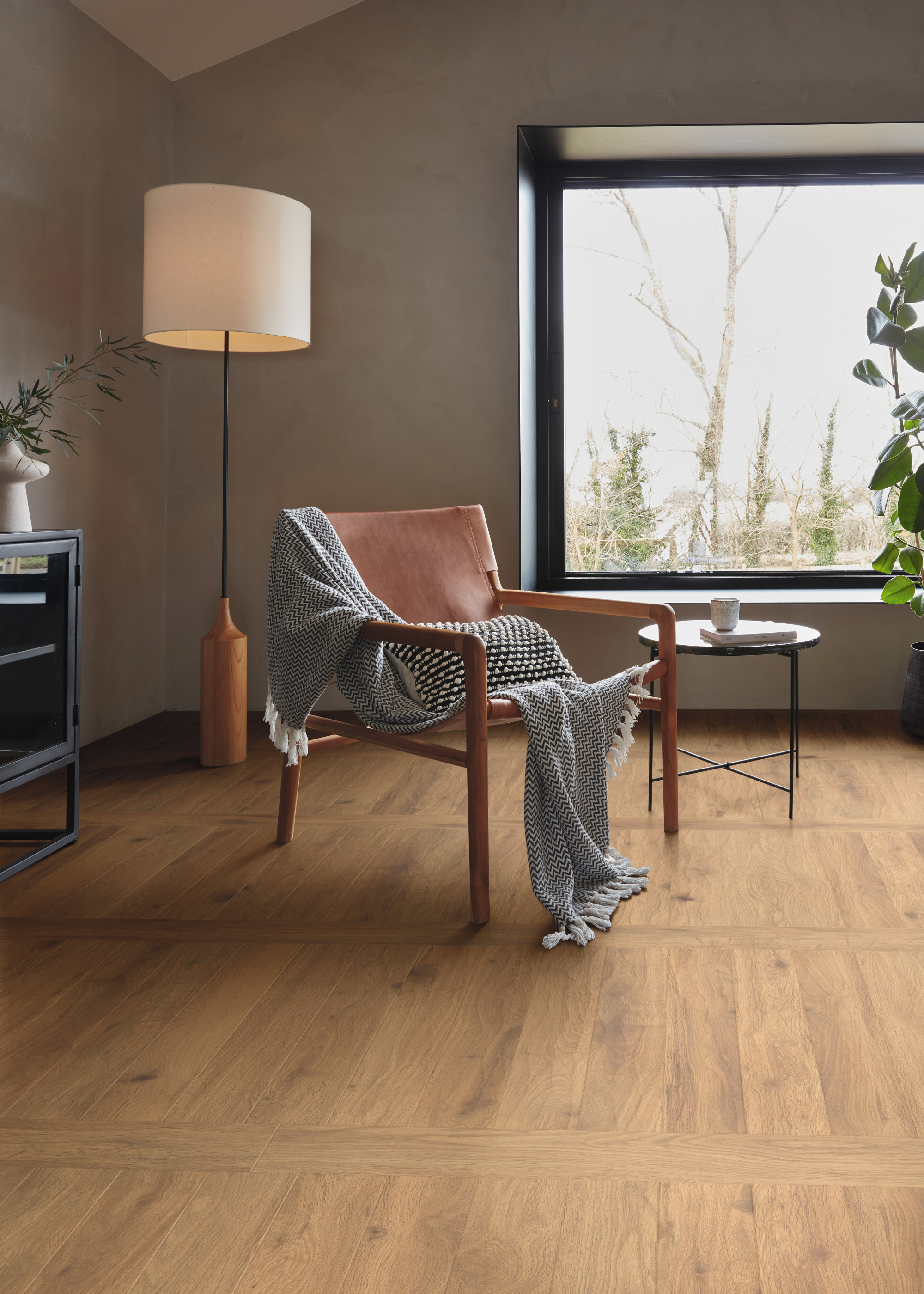 A warm living room with wood flooring, a chair with a throw and a pillow, a standing floor lamp, a side table with a cup, a potted plant and a large window