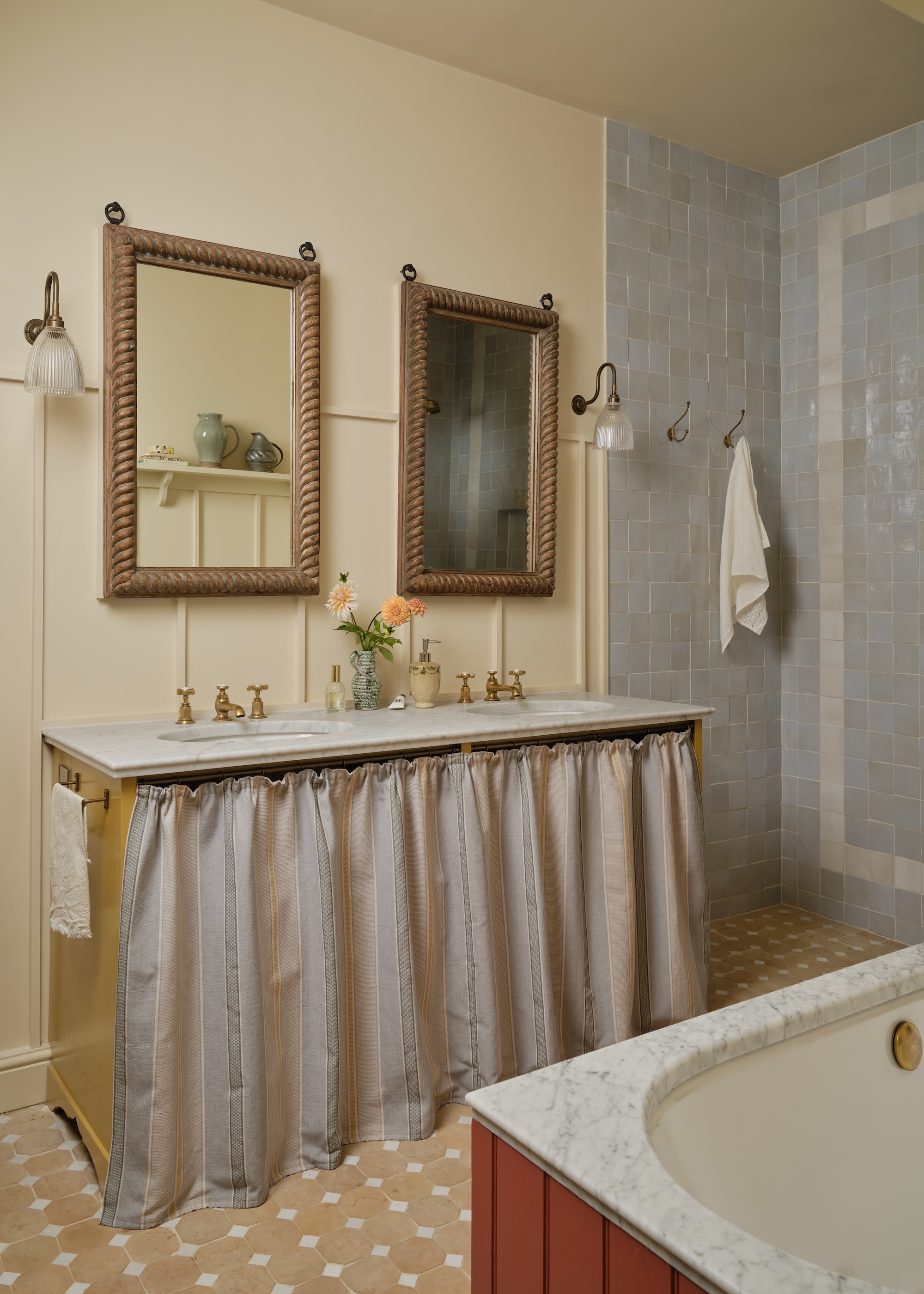 a bathroom showing a double vanity basin with a striped skirt, timber mirrors, wall lamps and walk in shower with gently textured tiles in pale blue