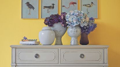 A dresser styled with flower vases in front of a yellow wall with bird art 