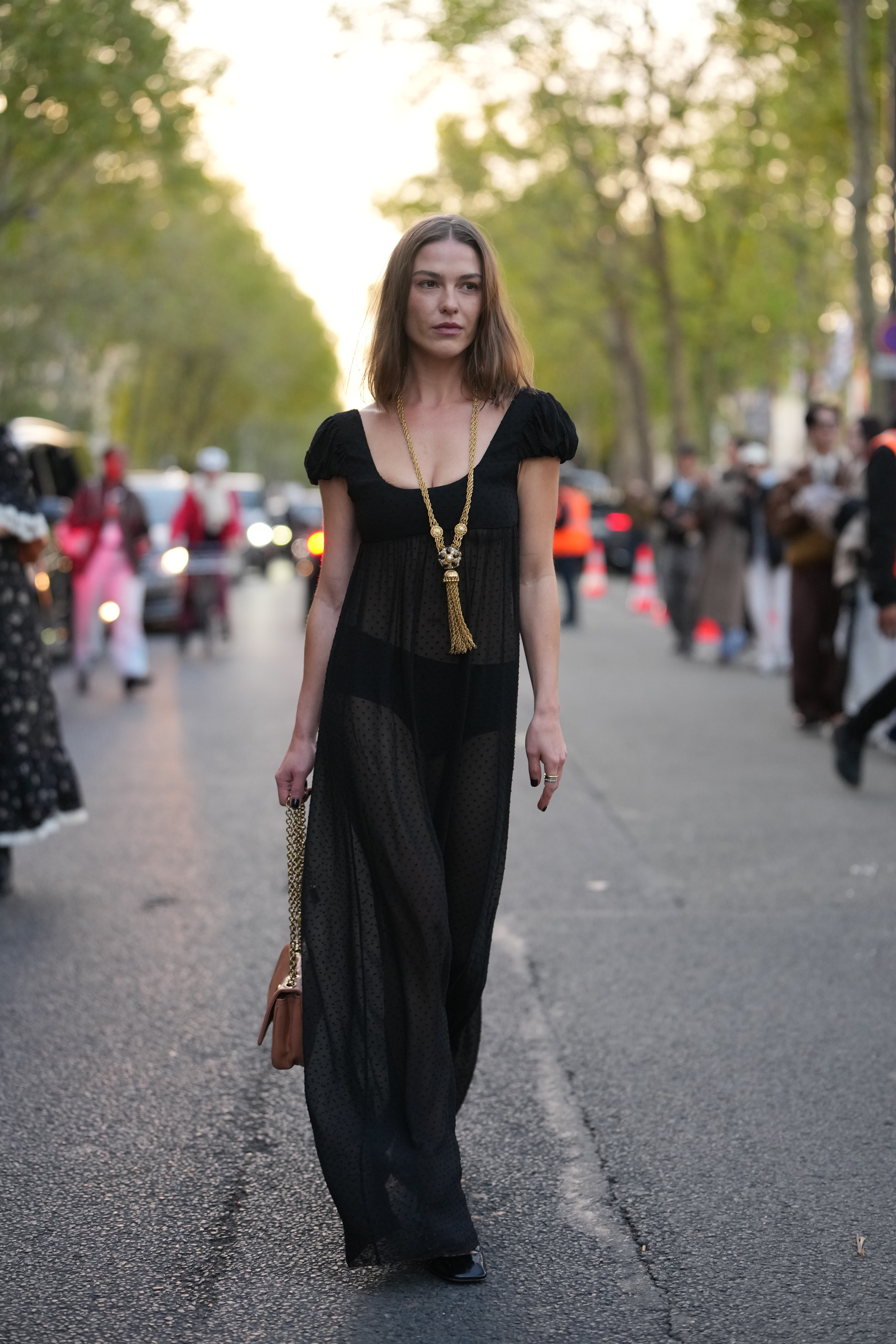 A guest wears shoulder-length straight brown hair with a center part, a long gold tasseled necklace with braided chain and ornamental knot, gold rings, a tan leather shoulder bag with gold chain strap and round gold clasp, a sheer black polka-dot chiffon maxi dress with puff short sleeves, a low scoop neckline, an empire seam and visible black undergarment briefs underneath, black patent leather loafers shoes with gold chain hardware and low block heels, outside Chloe, during Paris Fashion Week