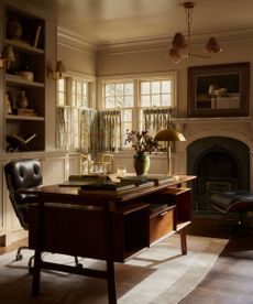 Cozy home office with dark wood desk, leather chair, fireplace and windows with floral cafe curtains to the right, bookshelf behind desk