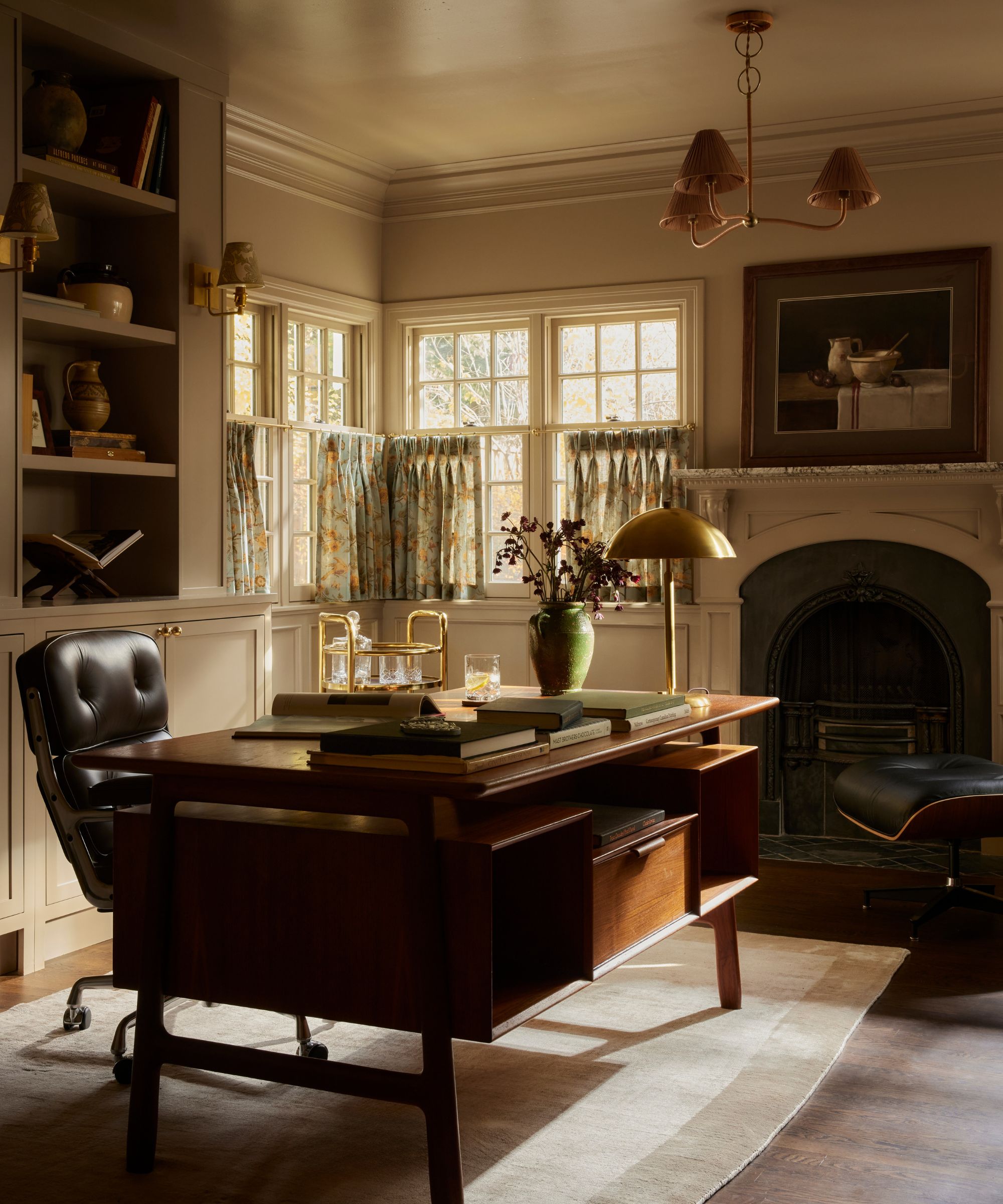 Cozy home office with dark wood desk, leather chair, fireplace and windows with floral cafe curtains to the right, bookshelf behind desk