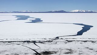 A frozen lake landscape with large cracks in the ice showing the deep blue water beneath. There are snow-capped mountains in the distance.