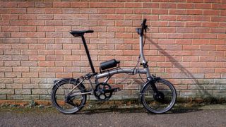 A silver Brompton T-Line, converted with the Cytronex e-bike conversion kit, leans against a brick wall